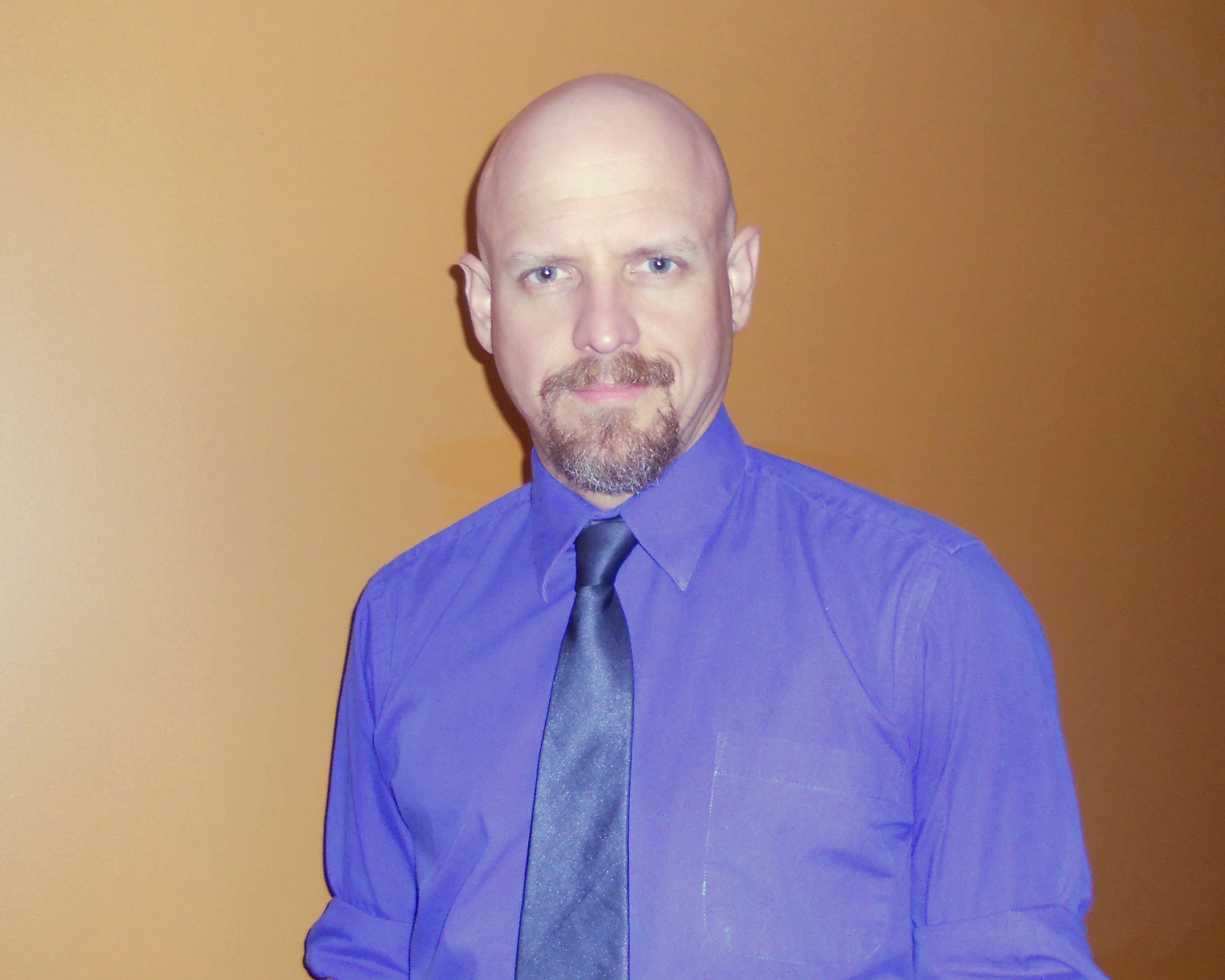 Portrait of Matthew Daniel Mason wearing a blue shirt and dark tie against an orange wall